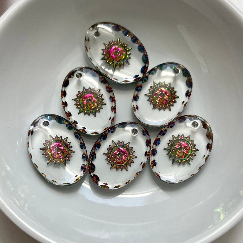 May include: Oval-shaped clear glass beads with a sunburst design in iridescent pink, yellow, and green. Each bead has a small hole at the top for stringing. The edges are decorated with small, colorful dots. These beads are ideal for jewelry making.