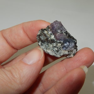 May include: A rough, dark gray rock with a purple and blue crystal embedded in it. The crystal is partially exposed and has a sharp, angular shape.