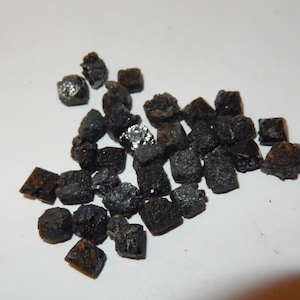 May include: A collection of small, dark, rough-textured, black crystals. The crystals are scattered across a white surface, with some catching the light and appearing shiny. The overall appearance suggests a mineral or gemstone.