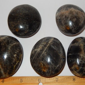 May include: Five smooth, polished black obsidian stones with a silvery sheen. The stones are oval shaped and have a natural, organic look. They are arranged on a white surface with a ruler for scale.
