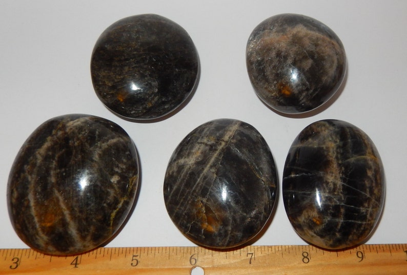 May include: Five smooth, polished black obsidian stones with a silvery sheen. The stones are oval shaped and have a natural, organic look. They are arranged on a white surface with a ruler for scale.