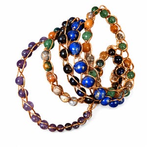 Könnte beinhalten: Eine Sammlung von drahtgewickelten Armbändern mit runden Edelsteinperlen in verschiedenen Farben wie Violett, Blau, Grün, Orange und Schwarz. Der Metalldraht ist um jede einzelne Perle gewickelt und bildet eine durchgehende kreisförmige Struktur.