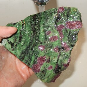 May include: A large, rough, green and pink gemstone with black veins. The stone is wet and appears to be under running water.