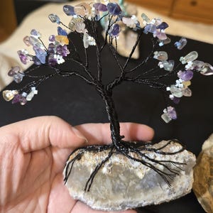 Può includere: Una scultura artigianale ad albero in filo di ferro con tronco e rami neri, adornata con schegge di pietre preziose colorate. L'albero è radicato in una base di pietra naturale, creando un pezzo decorativo unico.