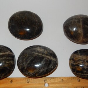 May include: Five smooth, polished black and brown stones, each about 2 inches in diameter, arranged on a white surface with a wooden ruler for scale.