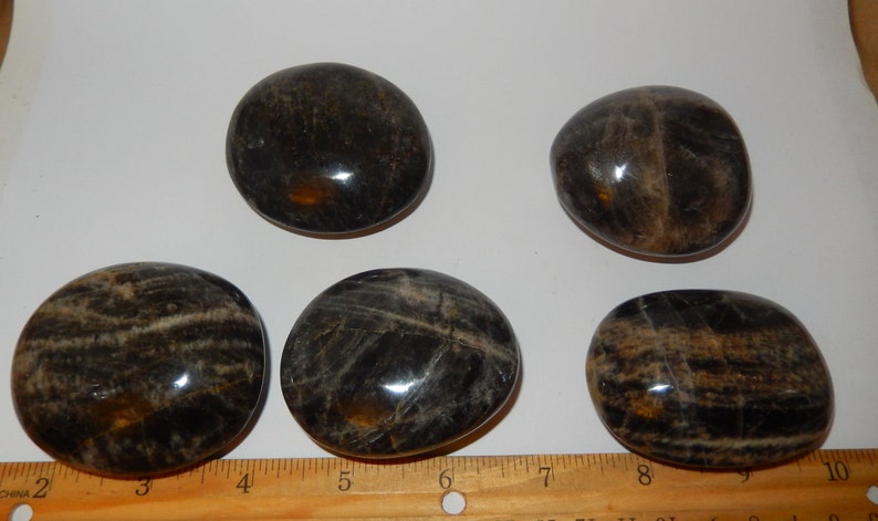 May include: Five smooth, polished black and brown stones, each about 2 inches in diameter, arranged on a white surface with a wooden ruler for scale.