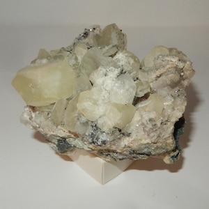 May include: A cluster of pale yellow, translucent crystals on a rough, gray and brown rock matrix. The crystals are clustered together in a natural formation, with some appearing to be partially embedded in the rock.