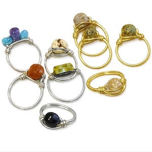 Könnte beinhalten: Eine Sammlung von drahtgewickelten Ringen mit verschiedenen Edelsteinen. Das Set enthält silber- und goldfarbene Metallringe, die mit polierten Steinen in Farben wie Violett, Blau, Orange, Grün, Schwarz und klaren Perlen verziert sind.