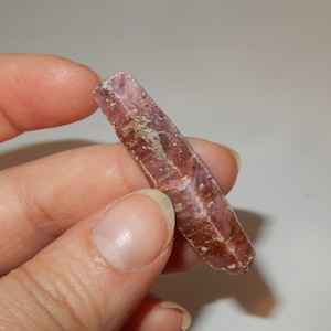 May include: A deep red, faceted, elongated crystal with a rough texture. The crystal is held in a hand.