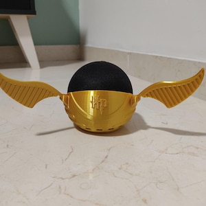 May include: A gold-colored 3D printed  'Golden Snitch' inspired cover for a smart speaker. The cover has a black speaker inside and two gold wings on either side. The cover has the letters 'HP' printed on the front.