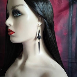 May include: A black earring with a flower-like design and a long, pointed dangle is worn by a mannequin. The earring has a small black bead and a gold-colored hook. The mannequin has long black hair and red lipstick.