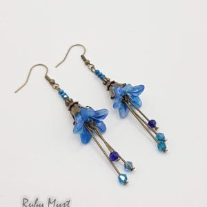 May include: A pair of handmade earrings featuring blue flower-shaped pendants. The earrings have bronze-colored hooks and accents, with blue and purple beads. The earrings are designed with a floral theme and are suitable for everyday wear.