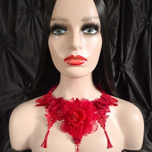 May include: A red lace necklace with a large rose in the center and tassel accents. The necklace is displayed on a mannequin with long black hair and red lipstick. The background is a dark, textured fabric.