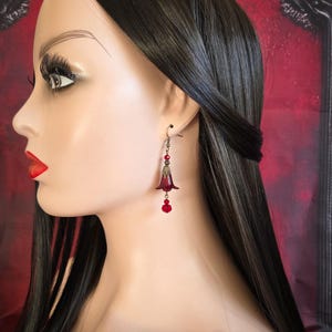 May include: A pair of dark red bell-shaped earrings with a dangling red bead. The earrings are attached to a mannequin's ear. The earrings have a vintage, gothic aesthetic.