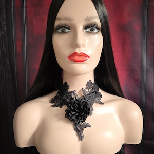 May include: Black lace choker necklace with a large black flower accent. The choker is made of delicate lace and features a floral design. The flower is made of fabric and has a ruffled texture.