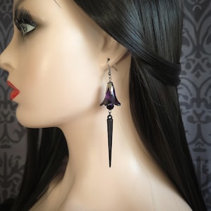 May include: A single earring with a dark purple flower-shaped pendant and a black, elongated spike. The earring hangs from a silver-coloured hook. The jewellery is displayed on a mannequin.
