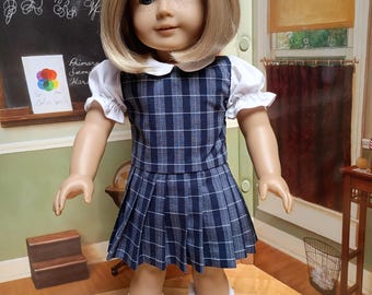 May include: A doll wearing a blue and white plaid dress with a white collared shirt and blue shoes. The doll has blonde hair and is standing in front of a chalkboard with the text "September 9, 1904" and "Penmanship".
