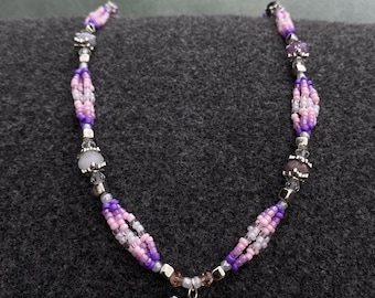 Puede incluir: Un collar de cuentas con un colgante de caballo plateado. El collar presenta un patrón de cuentas moradas, rosas y blancas. Las cuentas están ensartadas con metal color plata. El colgante de caballo es un diseño detallado en tres dimensiones.