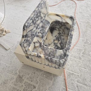 May include: A marble sink with a rectangular basin and a backsplash. The sink features a white and gray veined pattern. The base is a light beige color. The sink is likely for a bathroom or kitchen.