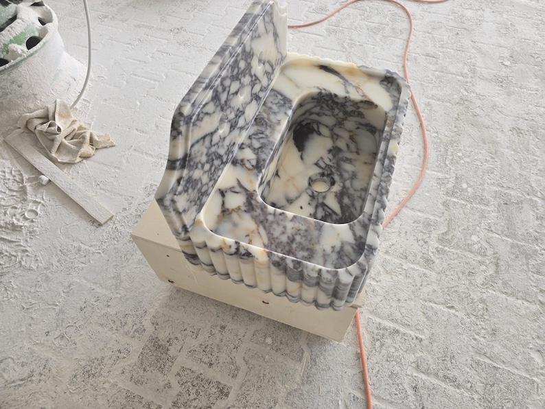 May include: A marble sink with a rectangular basin and a backsplash. The sink features a white and gray veined pattern. The base is a light beige color. The sink is likely for a bathroom or kitchen.