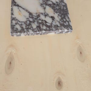 May include: A square marble tile with a white and gray pattern, resting on a light-colored wooden surface. The marble has a polished finish, and the wood grain is visible.
