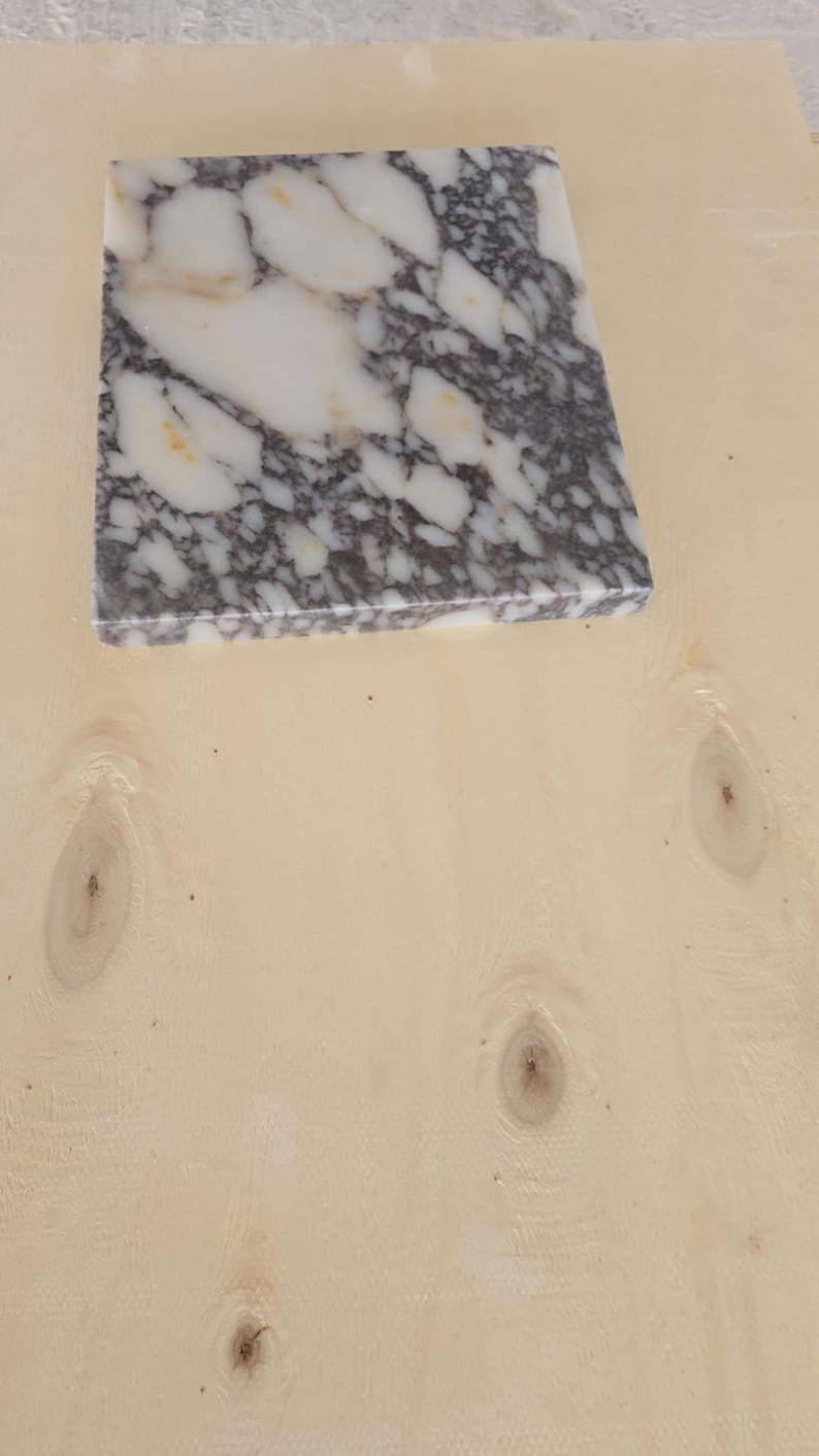 May include: A square marble tile with a white and gray pattern, resting on a light-colored wooden surface. The marble has a polished finish, and the wood grain is visible.
