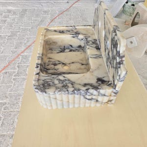 May include: A rectangular marble sink with a built-in backsplash. The sink basin is white with gray veining. The front edge of the sink has a fluted design. The backsplash has three holes.
