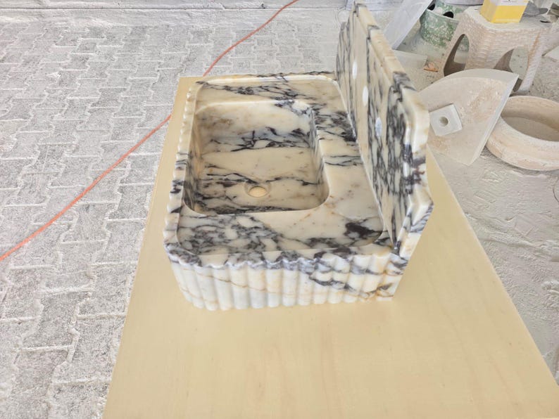 May include: A rectangular marble sink with a built-in backsplash. The sink basin is white with gray veining. The front edge of the sink has a fluted design. The backsplash has three holes.