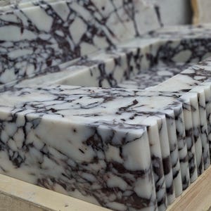 May include: Close-up of a marble countertop with a white base and dark purple veining. The edges are cut into a series of vertical ridges. The stone is displayed on a wooden pallet. This stone is suitable for kitchen or bathroom design.