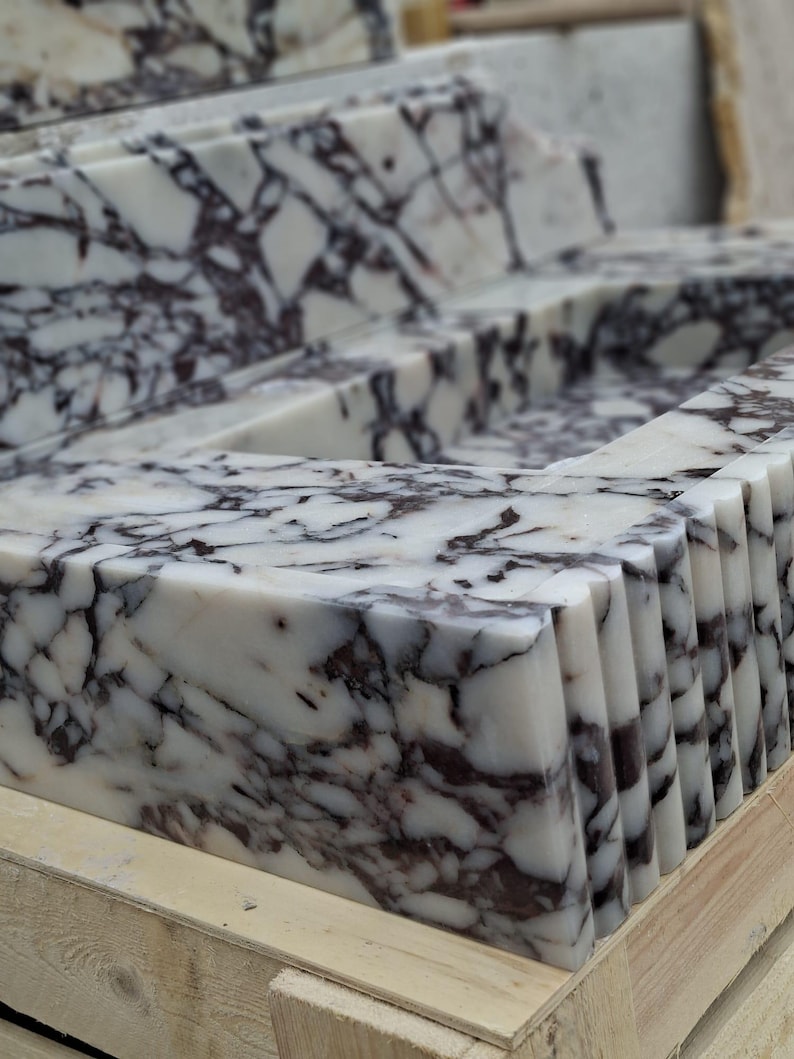 May include: Close-up of a marble countertop with a white base and dark purple veining. The edges are cut into a series of vertical ridges. The stone is displayed on a wooden pallet. This stone is suitable for kitchen or bathroom design.