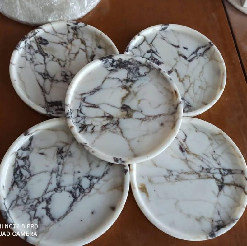 May include: Five white marble-patterned plates with dark gray and brown veining. The plates are round with a slightly raised edge. The plates are arranged in a circular pattern, showcasing the natural stone design.
