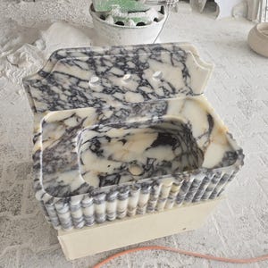 May include: A marble sink with a unique design. The sink features a white and gray veined pattern, a scalloped edge, and three faucet holes. The sink is set on a wooden base, showcasing a handcrafted aesthetic.