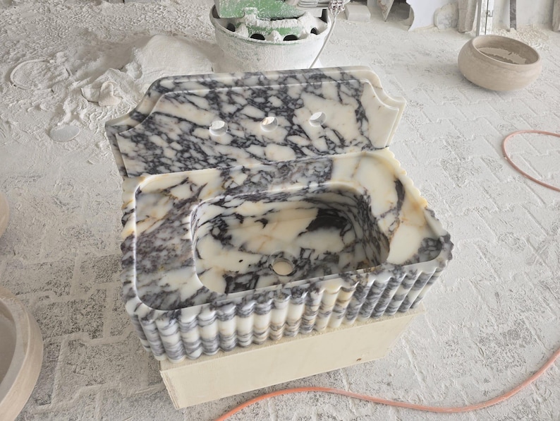 May include: A marble sink with a unique design. The sink features a white and gray veined pattern, a scalloped edge, and three faucet holes. The sink is set on a wooden base, showcasing a handcrafted aesthetic.