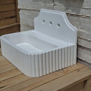 May include: A white, rectangular sink with a fluted front and a decorative back panel featuring three circular holes. The sink is made of a solid material and sits on a wooden surface.