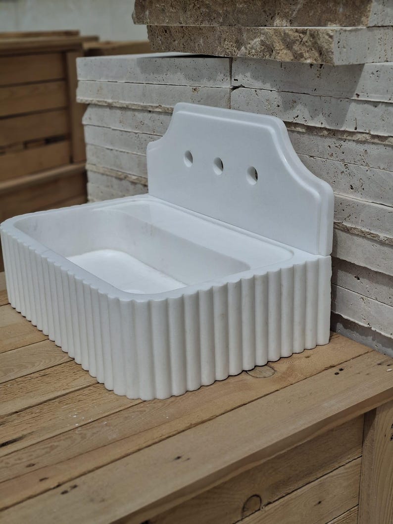 May include: A white, rectangular sink with a fluted front and a decorative back panel featuring three circular holes. The sink is made of a solid material and sits on a wooden surface.