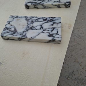 May include: Two rectangular marble tiles with a white base and intricate black veining. The tiles are placed on a light-colored wooden surface, showcasing the natural stone's pattern and texture. The tiles are likely for home decor or construction.