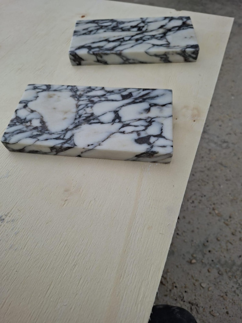 May include: Two rectangular marble tiles with a white base and intricate black veining. The tiles are placed on a light-colored wooden surface, showcasing the natural stone's pattern and texture. The tiles are likely for home decor or construction.