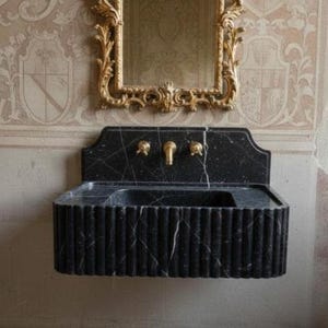 May include: A black marble sink with gold fixtures is mounted on a wall. The sink has a fluted design and is paired with a gold-framed mirror. The wall features ornate wallpaper and a small wooden shelf.