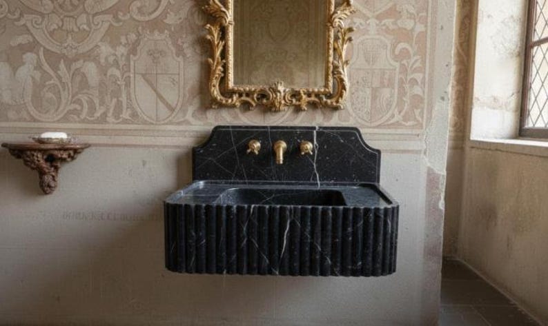 May include: A black marble sink with gold fixtures is mounted on a wall. The sink has a fluted design and is paired with a gold-framed mirror. The wall features ornate wallpaper and a small wooden shelf.