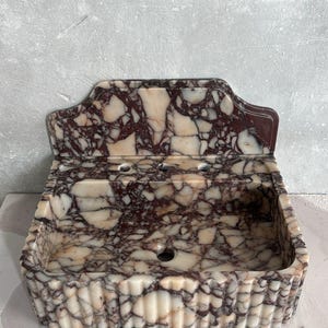 May include: A marble sink with a rectangular basin and a decorative backsplash. The sink features a pattern of burgundy and cream-colored veining. The front of the sink has a fluted design.