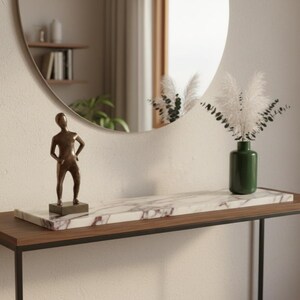May include: A modern console table with a dark wood top and black metal frame. A bronze statue, a marble tray, and a green vase with white plumes are displayed. A round mirror hangs on the wall.