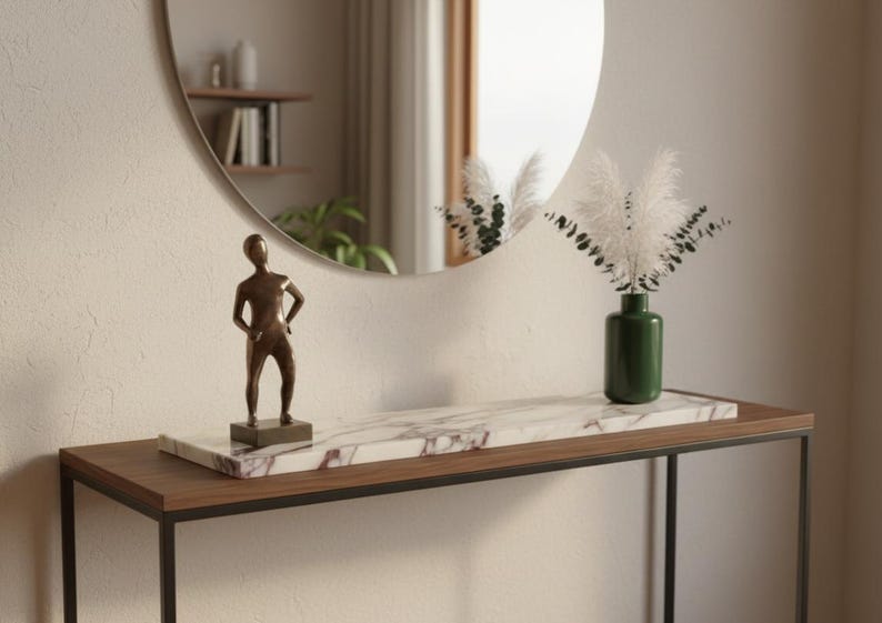 May include: A modern console table with a dark wood top and black metal frame. A bronze statue, a marble tray, and a green vase with white plumes are displayed. A round mirror hangs on the wall.