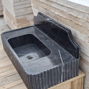 May include: A black marble sink with a rectangular basin and a fluted exterior. The sink has a built-in backsplash and is displayed on a wooden surface. The dark stone features white veining.