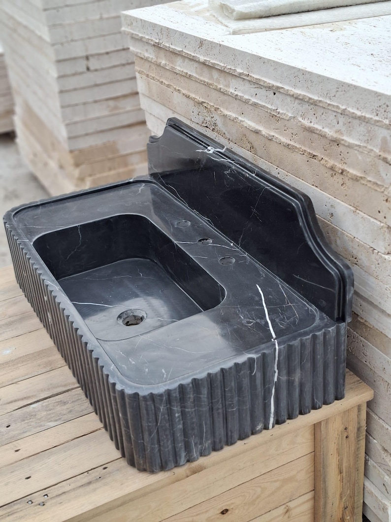 May include: A black marble sink with a rectangular basin and a fluted exterior. The sink has a built-in backsplash and is displayed on a wooden surface. The dark stone features white veining.