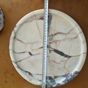 May include: A round marble tray with a cream and brown veined pattern. The tray is approximately 12 inches in diameter. A partial view of a matching marble object is visible on the left.