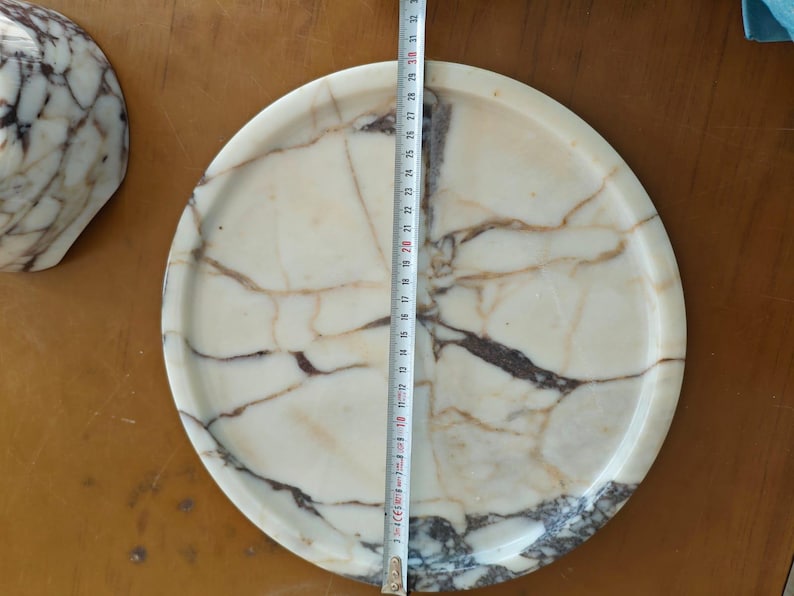 May include: A round marble tray with a cream and brown veined pattern. The tray is approximately 12 inches in diameter. A partial view of a matching marble object is visible on the left.