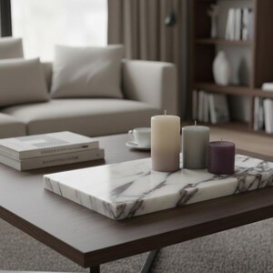 May include: A living room scene featuring a dark wood coffee table with a marble tray holding three pillar candles in cream, gray, and purple. Two books are stacked on the table, and a sofa is in the background.