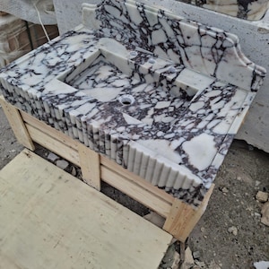 May include: A white marble bathroom sink with dark purple veining. The sink has a rectangular basin and a decorative backsplash. It is set on a wooden crate. The sink has a unique, fluted edge design.