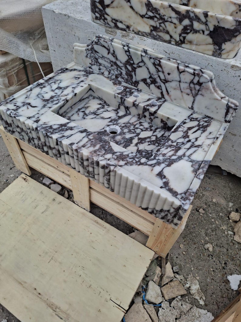 May include: A white marble bathroom sink with dark purple veining. The sink has a rectangular basin and a decorative backsplash. It is set on a wooden crate. The sink has a unique, fluted edge design.