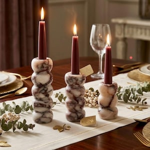 May include: Three stacked marble candle holders in varying heights, each featuring a cylindrical shape with rounded segments and a polished finish. Each holder contains a lit, slender, deep burgundy taper candle with a visible flame.